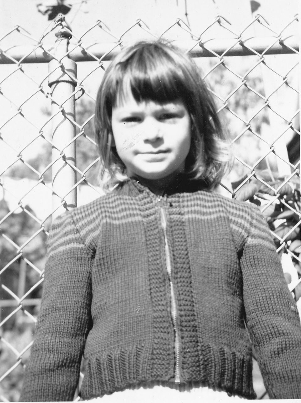 June Guralnick as a child in a sweater in front of a chain link fence - black and white photo