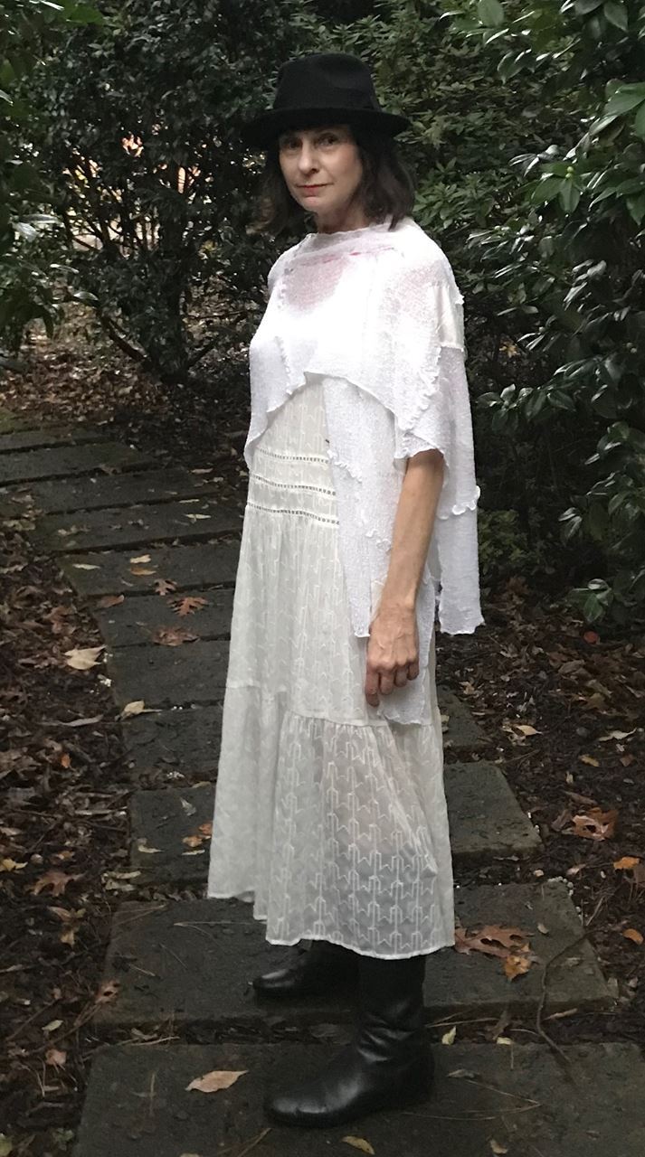 June Guralnick in a white dress with black boots and black hat standing outdoors on a stone path